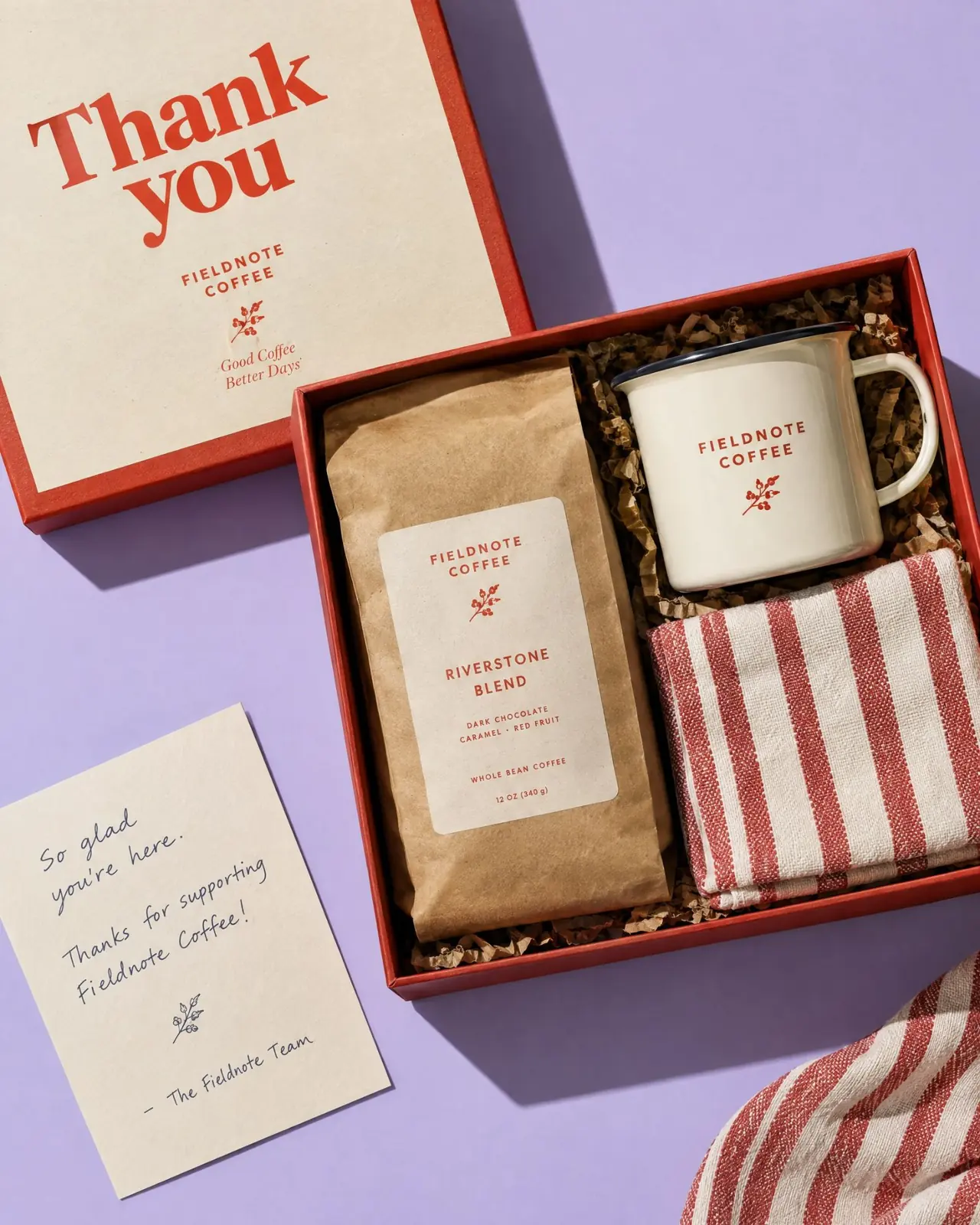 An open red Fieldnote Coffee gift box with a coffee bag, enamel mug and striped tea towel, beside a Thank you lid and a handwritten card
