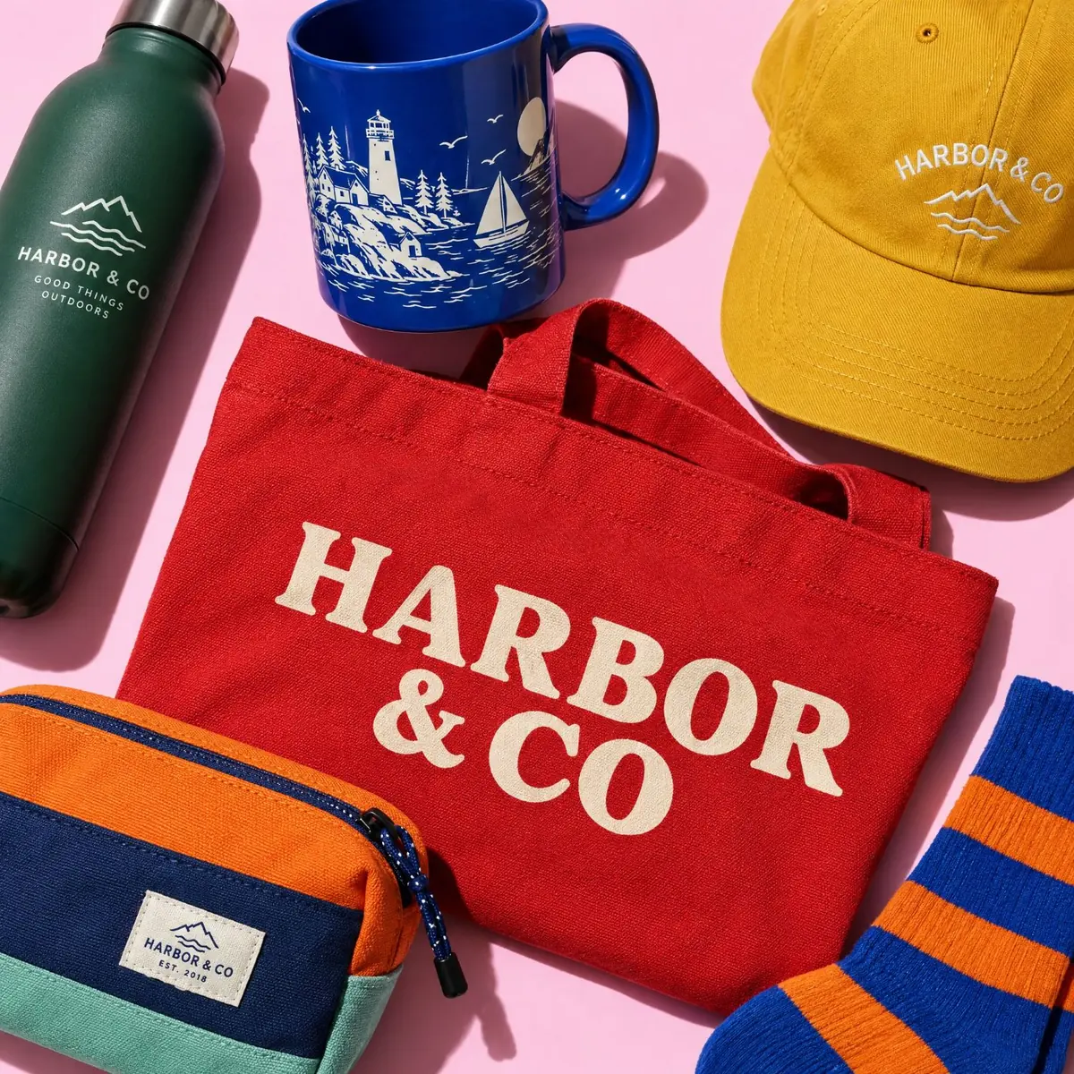 A red Harbor and Co tote bag with a green water bottle, a blue mug, a yellow cap, a zip pouch and striped socks on a pink background