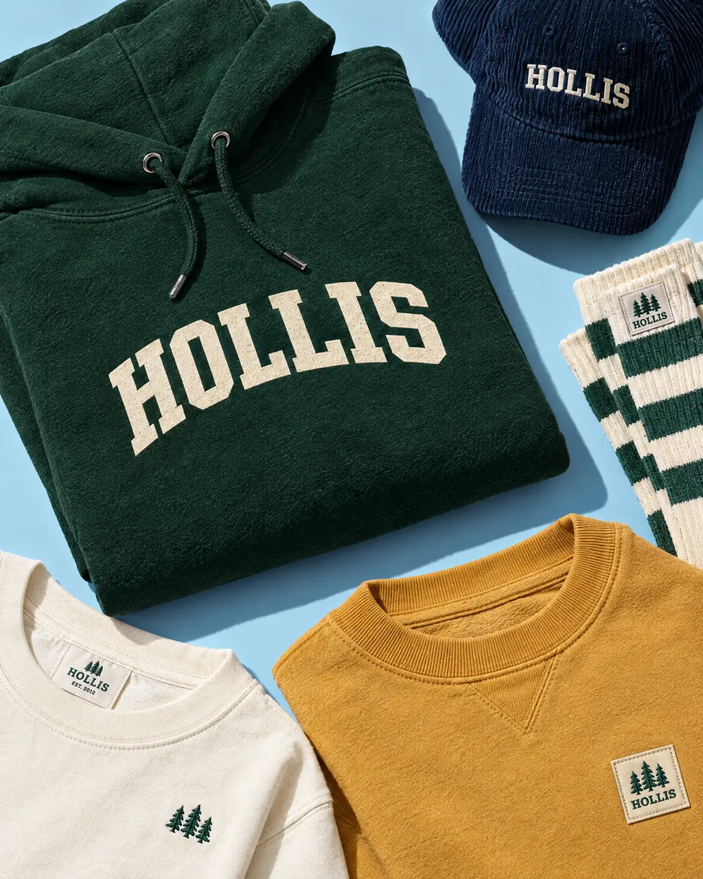 A green Hollis hoodie, a navy corduroy cap, striped socks, a cream t-shirt and a mustard sweatshirt laid flat on a blue background