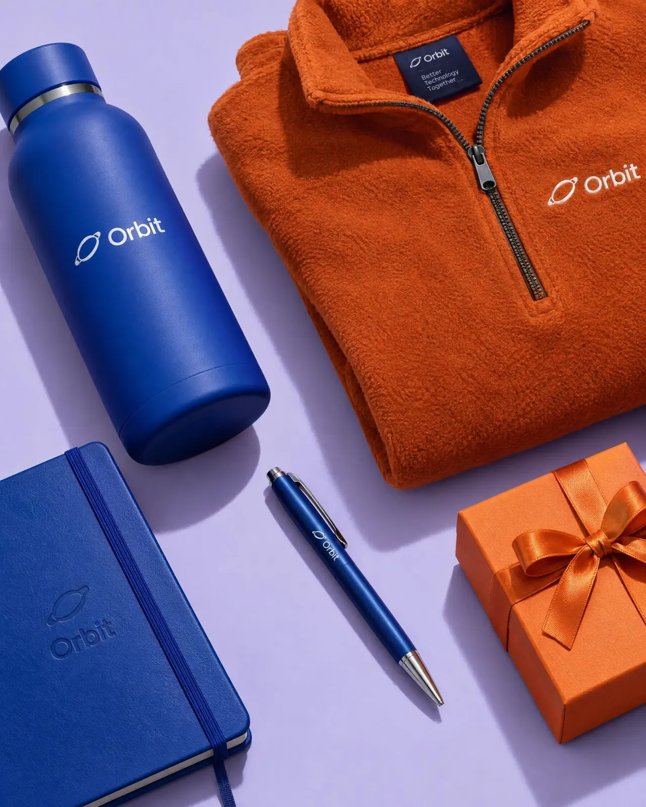 A blue Orbit water bottle, a folded orange quarter-zip fleece, a blue notebook and pen, and an orange gift box on a lilac background